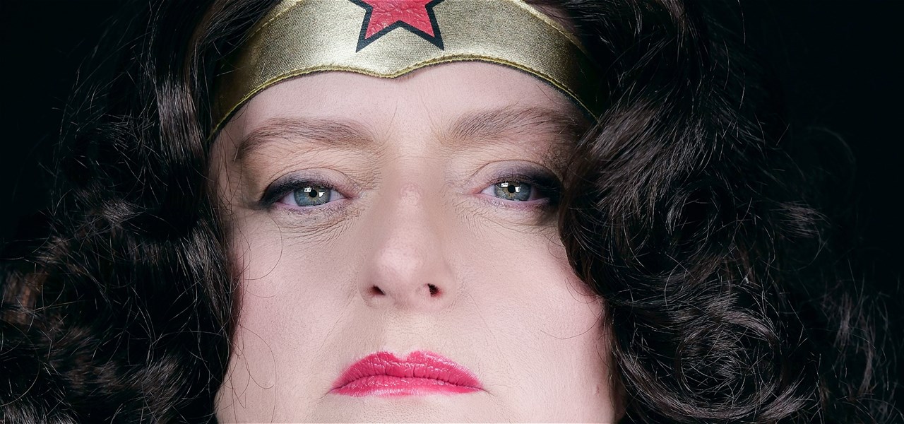 A white woman with dark eyes, curly dark brown hair is close up to the lens with a gold headband and a star looking seriously at you.