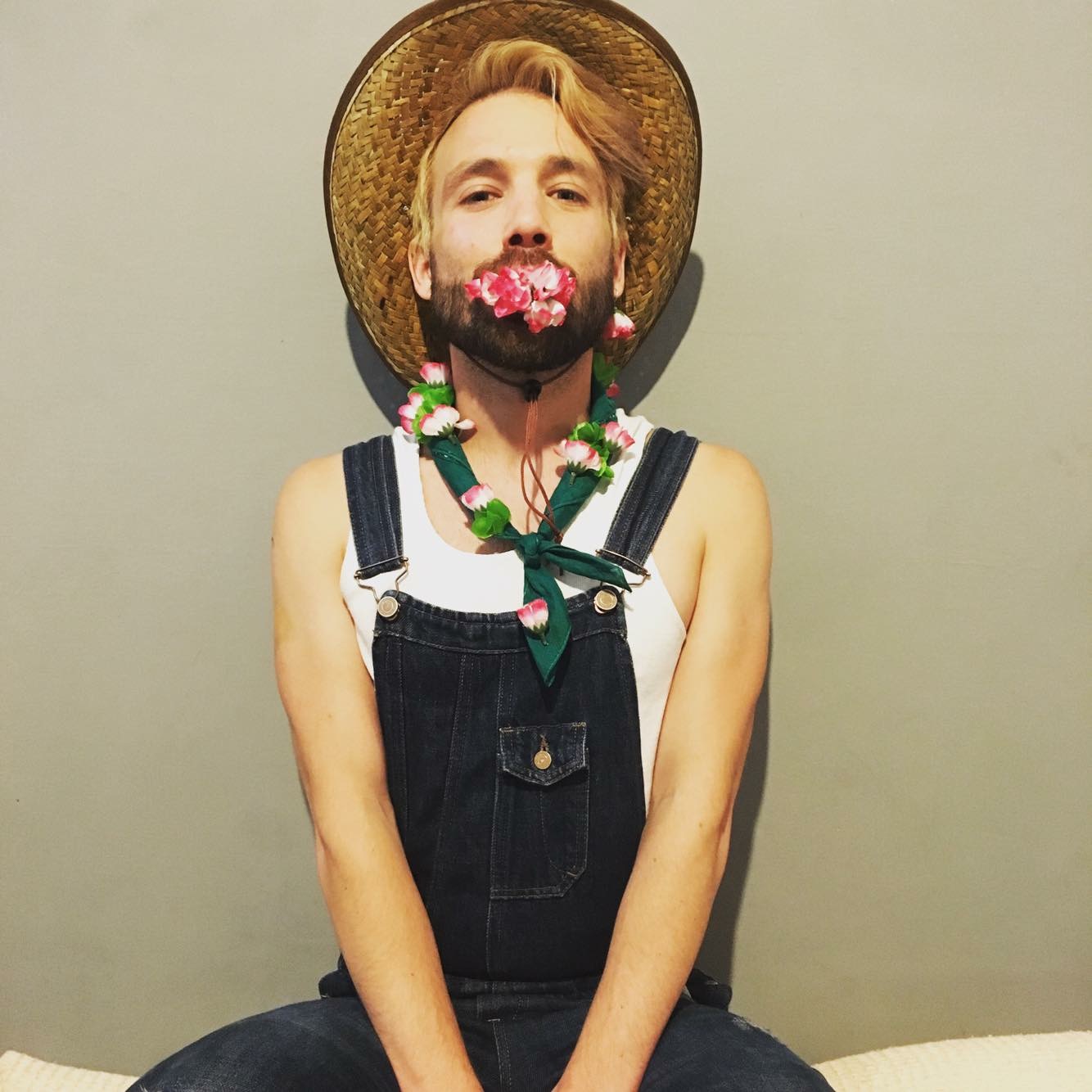 Portrait of Matt wearing overalls, a straw hat, a mouthful of flowers and more flowers decorating the overalls