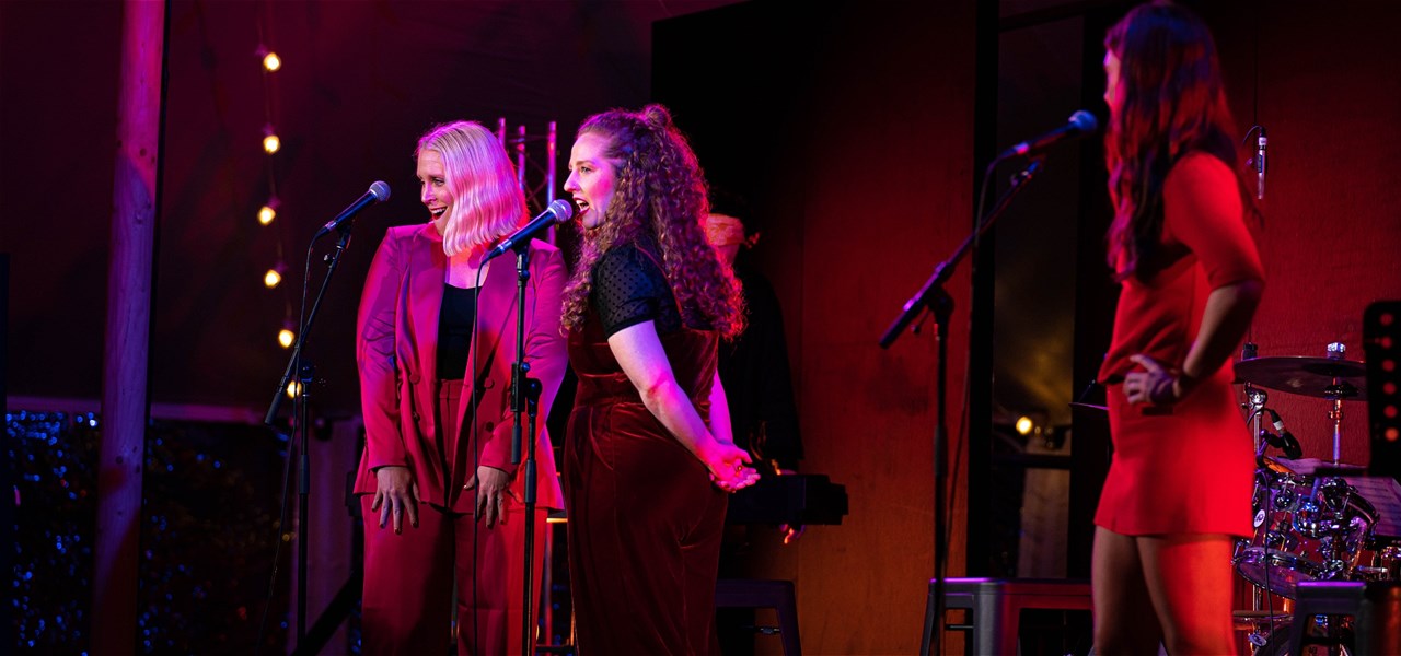 3 women dressed in colours of red and pink and all singing into microphones
