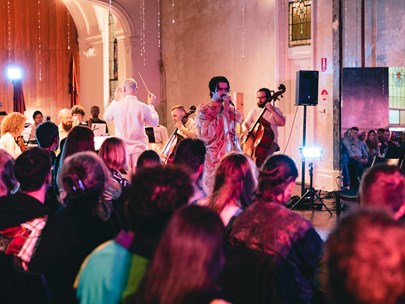 A crowd of people in a church space watch a performer in a flowing dress sing. There is an orchestra behind them. 