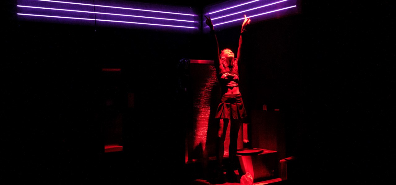 Person with arms outstretched standing on a dark stage with red lighting and mauve neons overhead