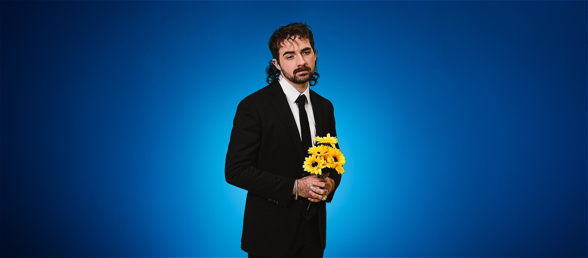 The lead performer stands in a black suit in front of a blue backdrop. He holds yellow flowers, and looks sadly into the distance