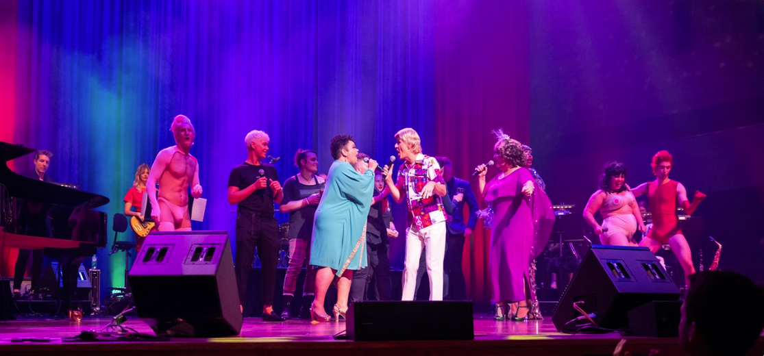 A crowd of singers, dancers, and musicians performing in colourful clothes against a rainbow curtained backdrop