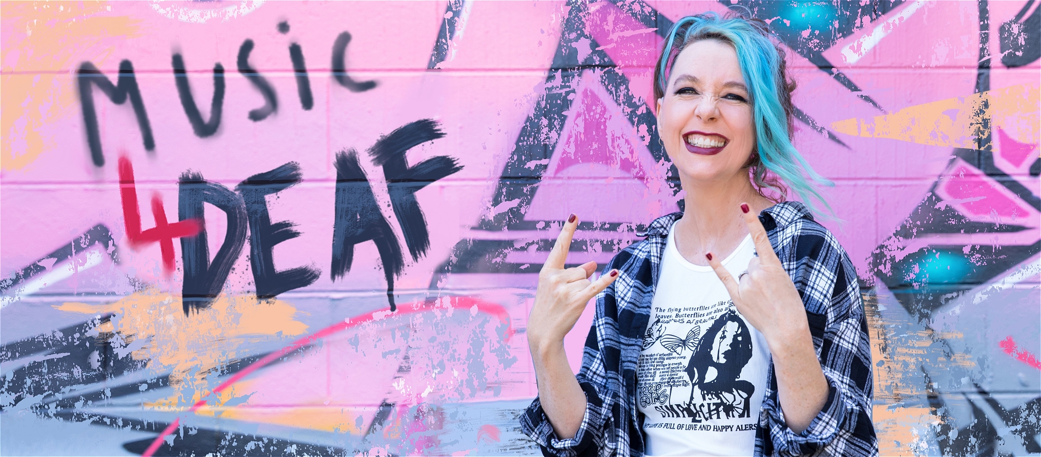 A person with blue hair smiling and standing in front of a colourful wall with text saying - Music 4 DEAF