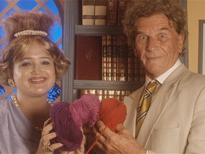 Two people in vintage dress face the camera smiling. They are both clutching balls of wool.
