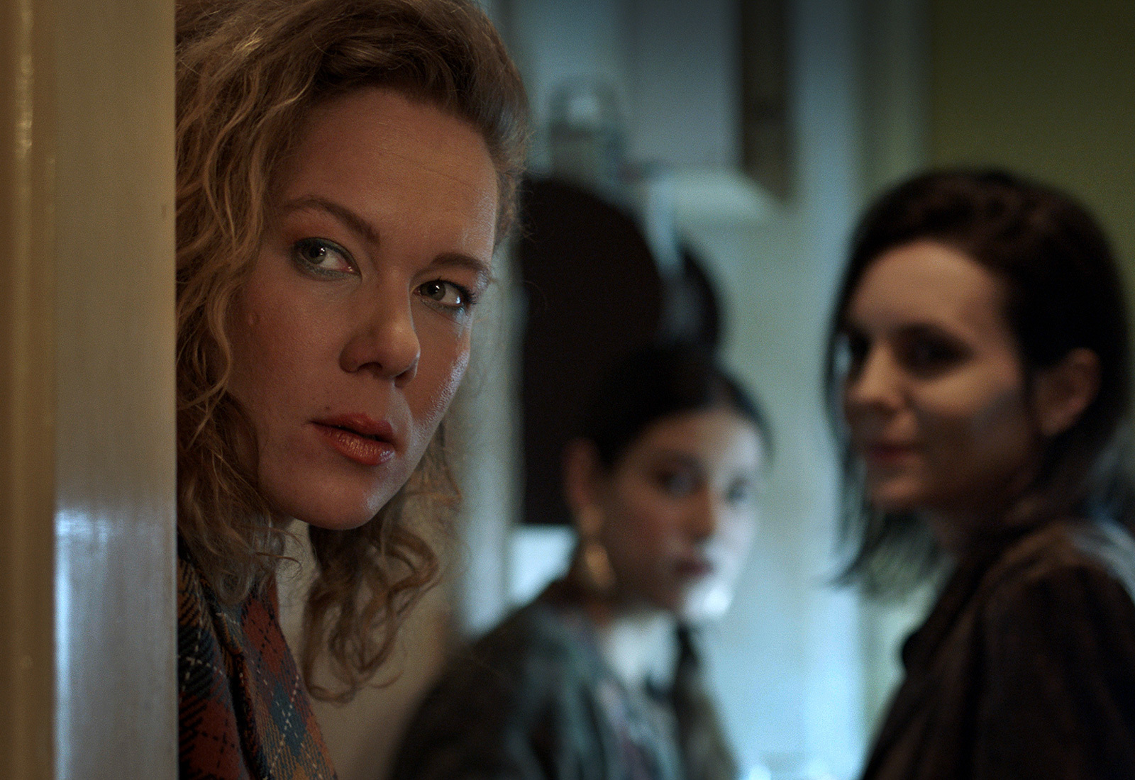 Three women in a home setting, with the one at the front peering towards the camera through a doorway