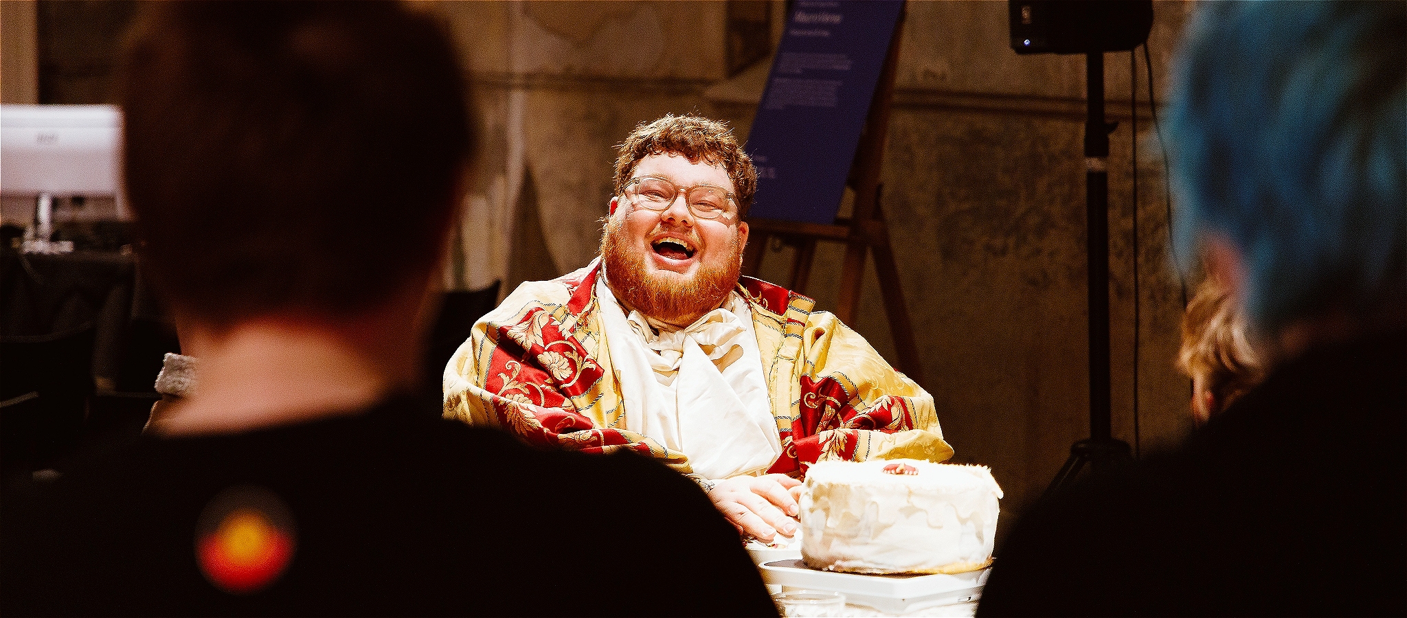 A person with short ginger hair and a beard wears glasses, smiling whilst sitting at a table infront of some artwork that looks like cake.