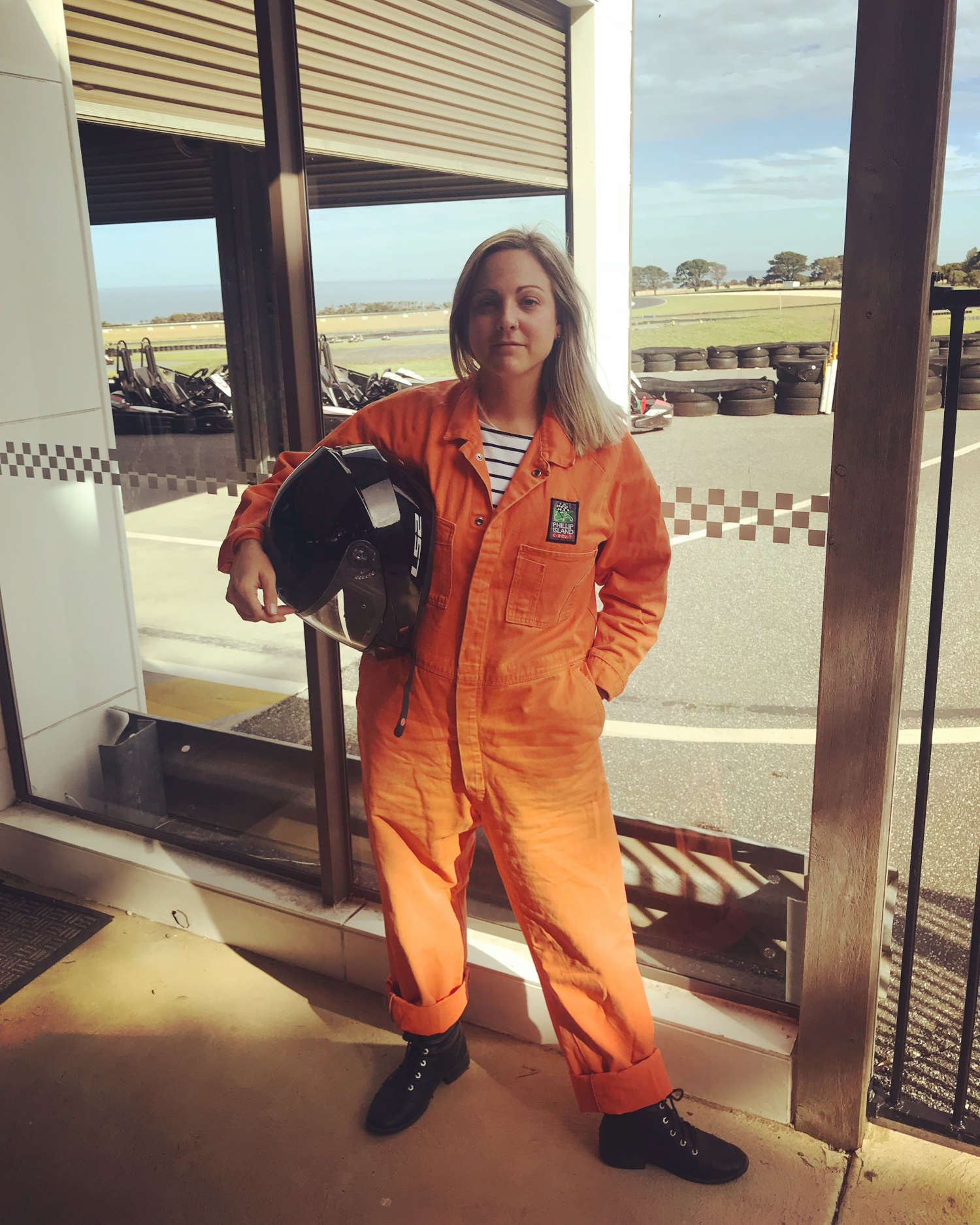 Photo of Flick dressed in orange overalls and holding a motorbike helmet, with a race track in the background