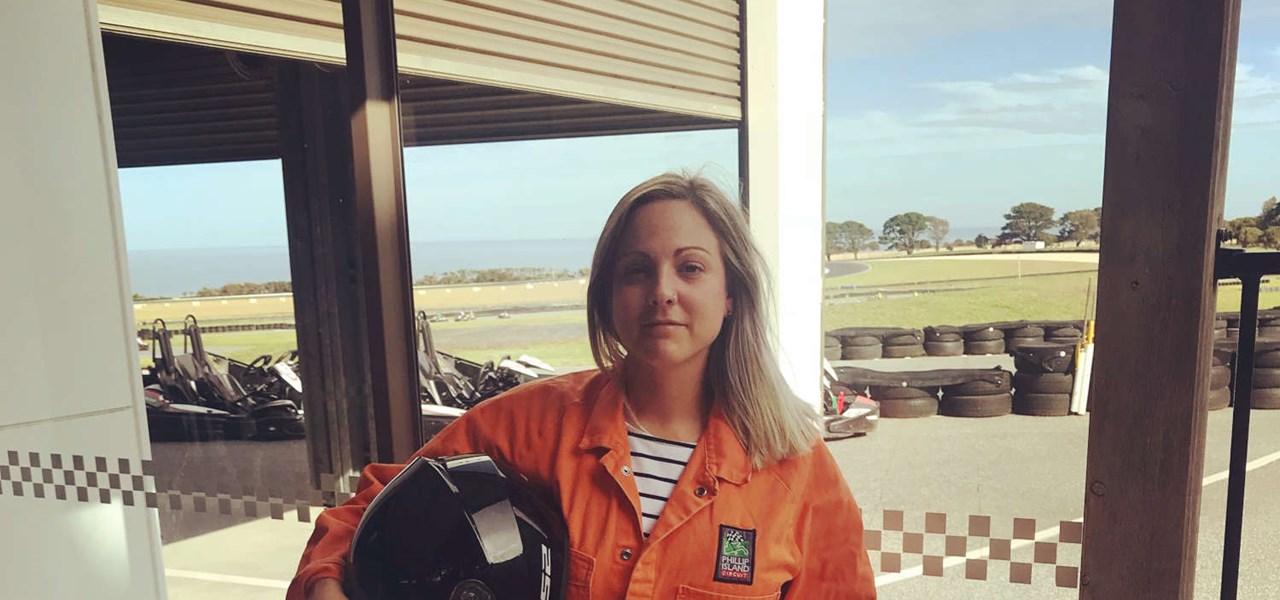 Photo of Flick dressed in orange overalls and holding a motorbike helmet, with a race track in the background