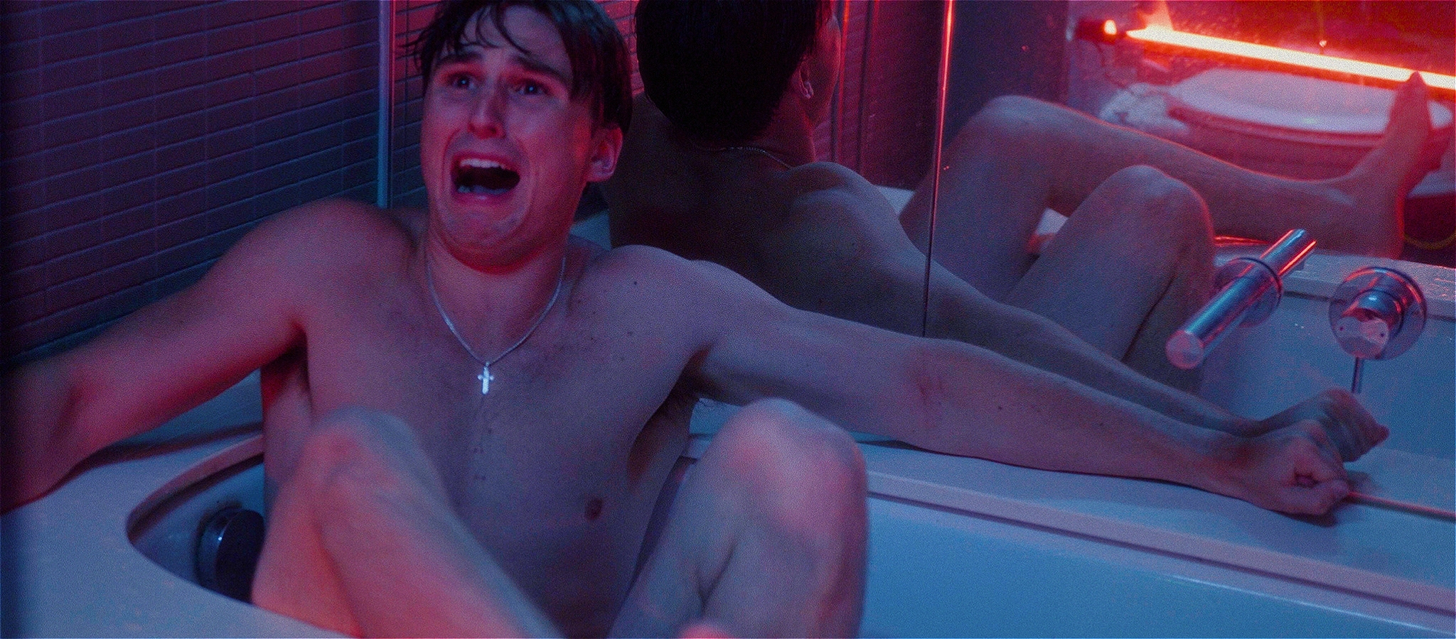 A naked person sitting in a bath with mirrors behind them looks scared looking at someone off camera who seems to be in the space.
