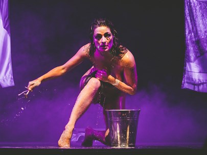 Headshot of Betty Grumble on stage with mauve lighting, kneeling behind a bucket