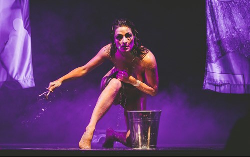 Headshot of Betty Grumble on stage with mauve lighting, kneeling behind a bucket