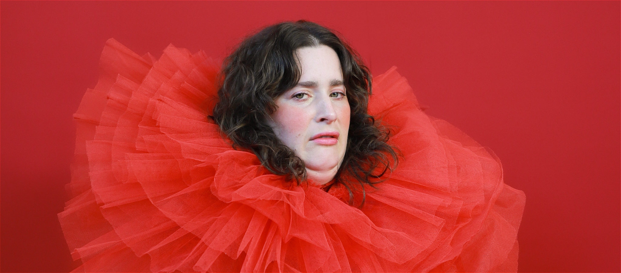 A woman with a ridiculously oversized red tulle neck ruffle stares into the camera against a red backdrop