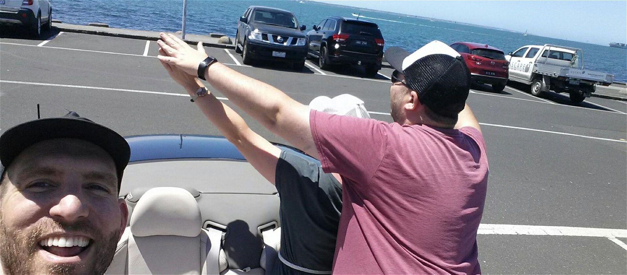 A bearded person smiling and taking a selfie with two people standing in a car, facing backwards and replicating the Titanic pose.