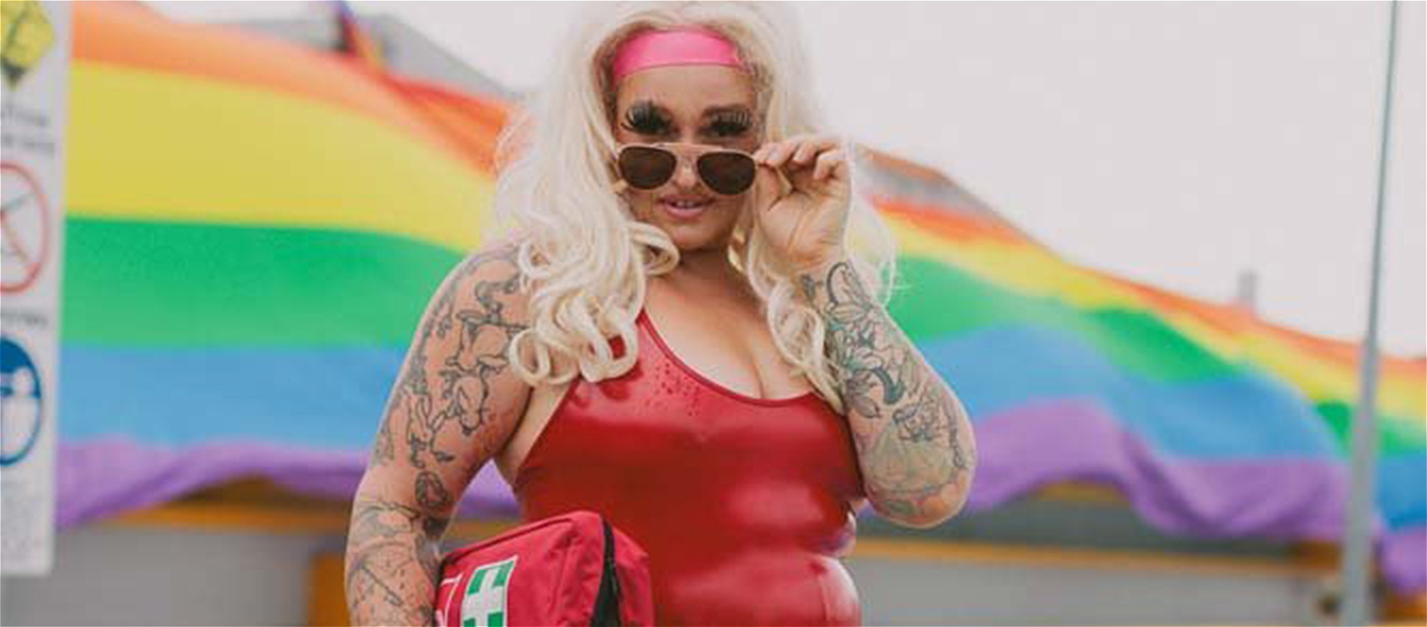 A performer dressed up in a lifeguard costume carrying a first aid kit poses looking over her sunglasses in front of a rainbow background.