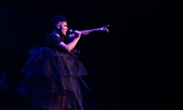 Sheldon Riley performing at Midsumma Festival 2020 in a large black dress, holding a mic in one hand, the other stretching towards the audience