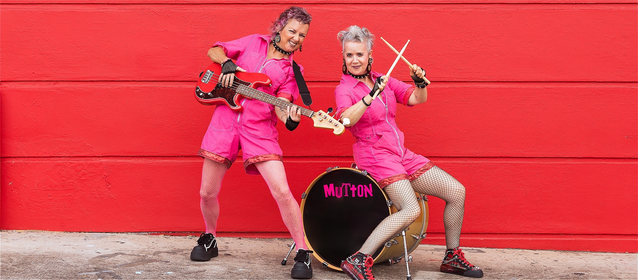 Two women in hot pink short overalls posing with a bass and a drum kit, smiling