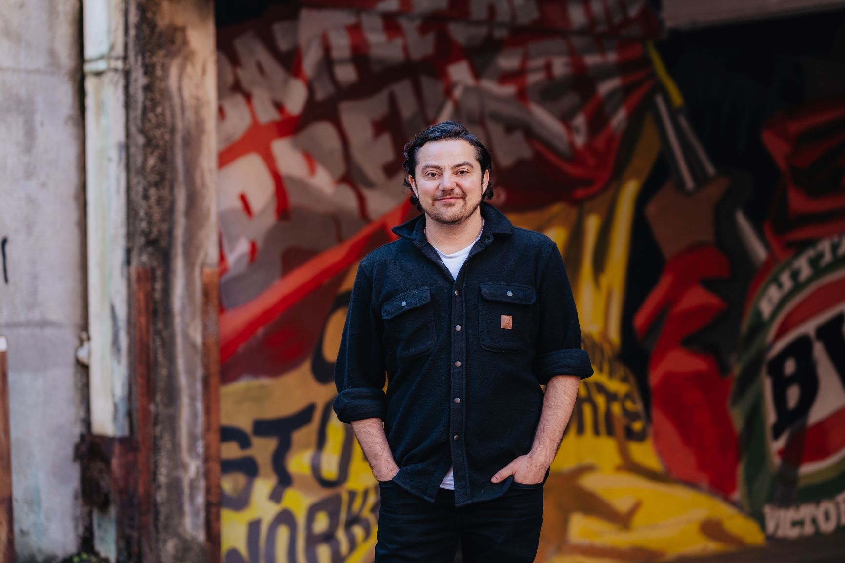 A person standing in a black shirt with their hands in their pockets smiling.