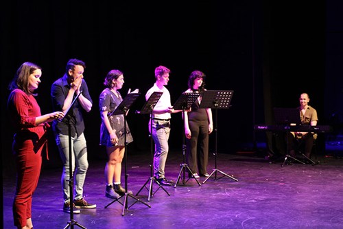 People standing on stage, one at a lectern and one sitting at a piano