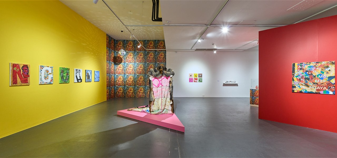 An empty large gallery space with art on a yellow wall on the left, a red one on the right and a stand-alone exhibit in the centre of the room