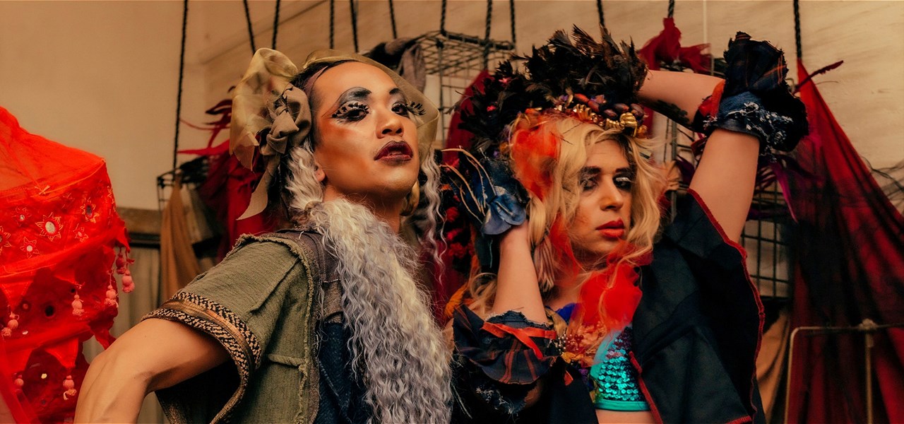 Two drag artists pose in front of a sewing studio, they both wear layered garments in different darker tones and textures.