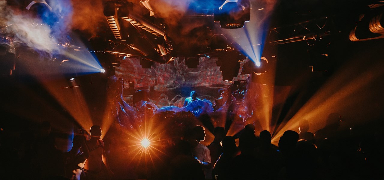 A group of people dancing amongst bright lights and in the background, a blue light shines brightly on the DJ booth.