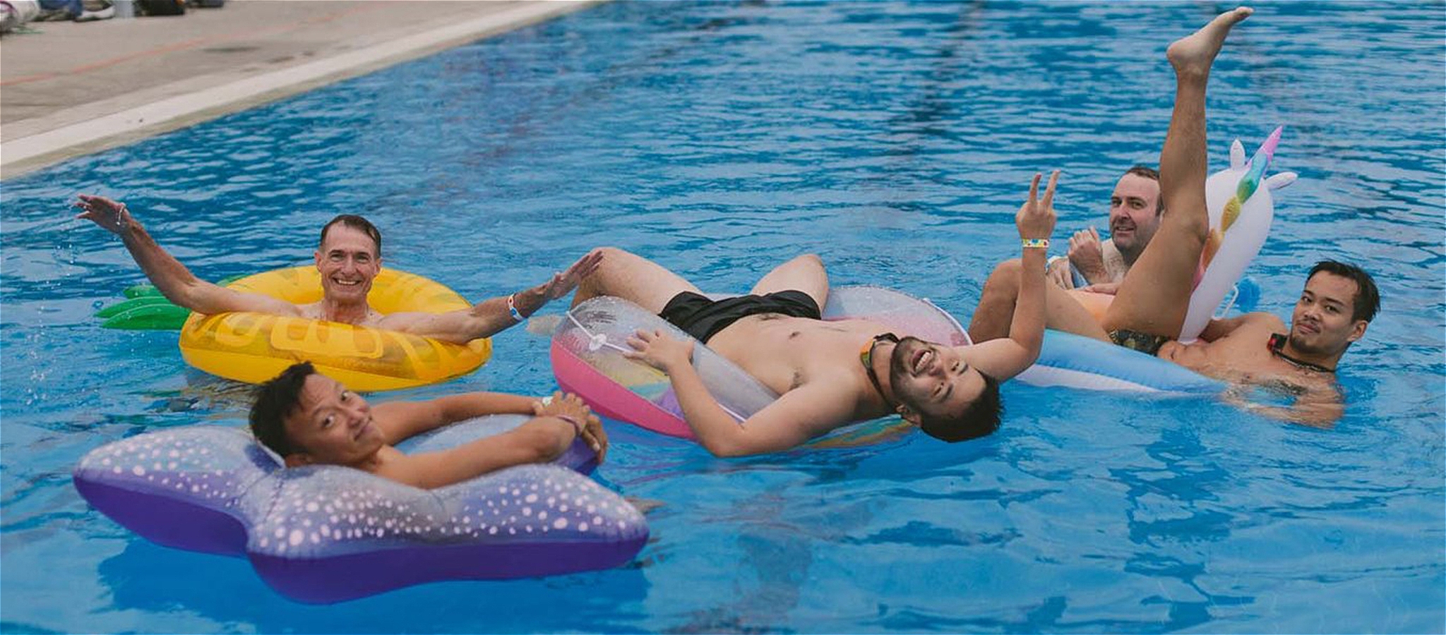 Five people smiling and posing while laying on top of fun floaties in the pool.