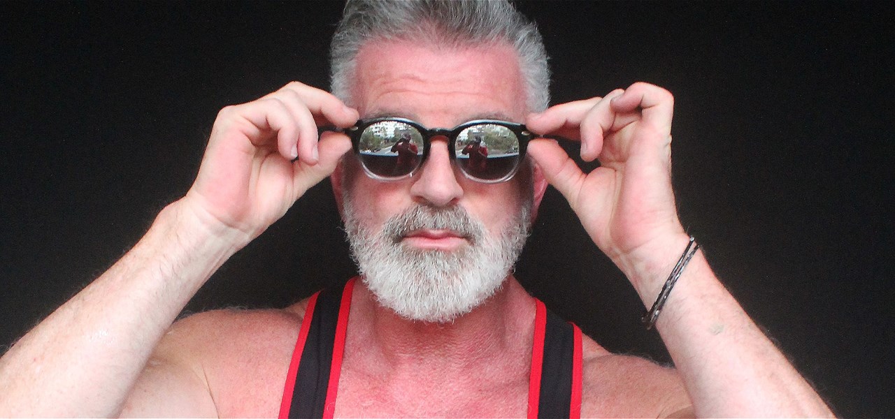 An older gay man in a singlet with hands up to hold his sunglasses