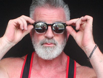 An older gay man in a singlet with hands up to hold his sunglasses