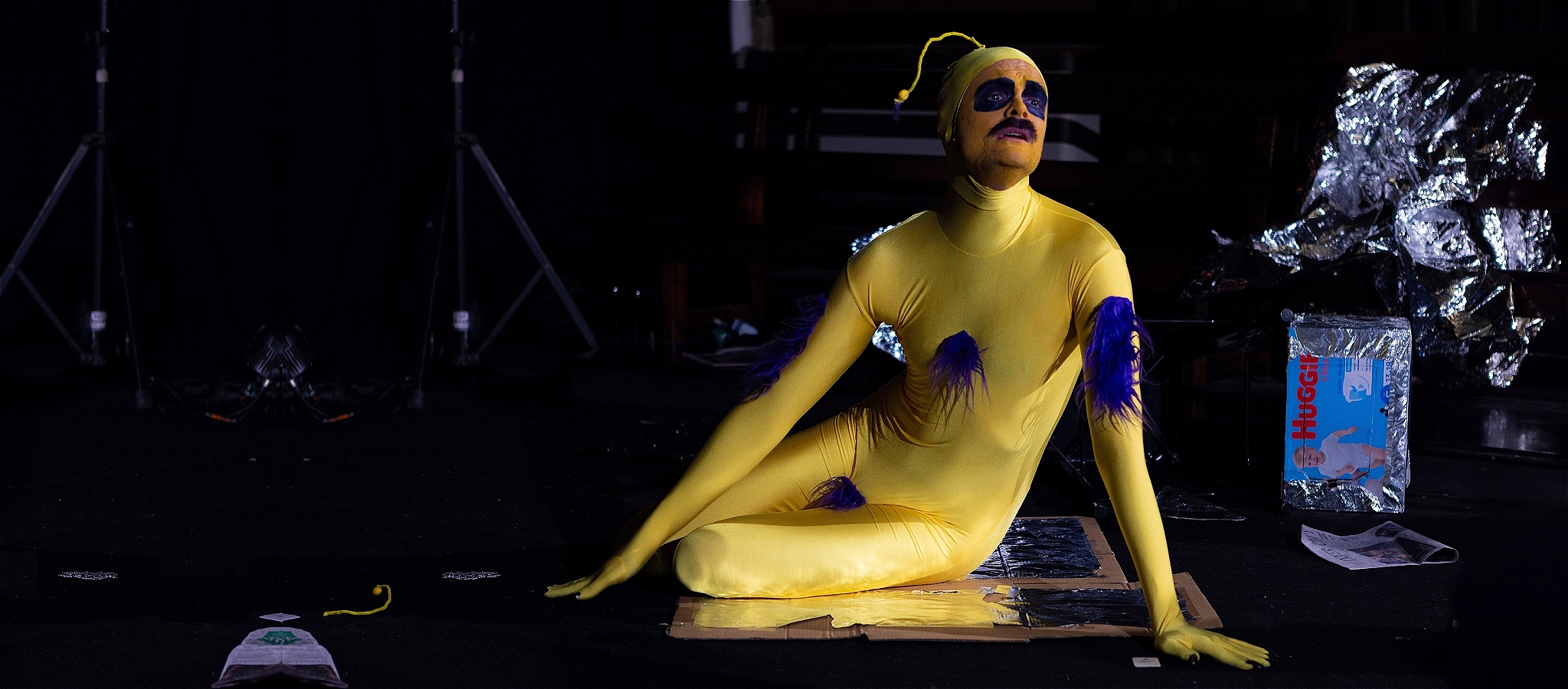 84BY, a yellow alien with dark eye makeup and purple fur patches, sits dramatically on the floor amid scattered debris and stage props.