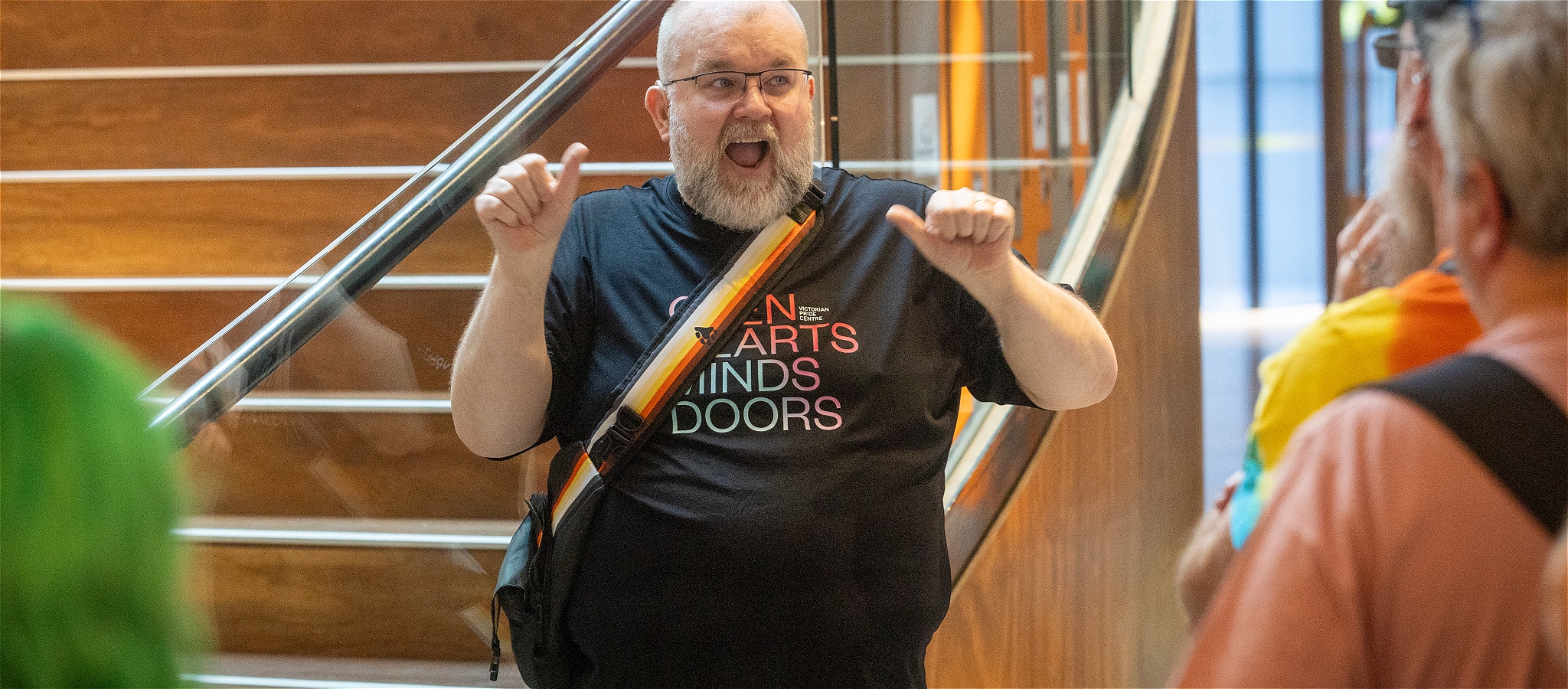 A person signing and smiling while standing in front of the staircase at the Victorian Pride Centre
