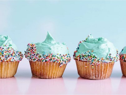 A row of cupcakes sitting on a light mauve surface, with aqua icing and hundreds & thousands around the rim