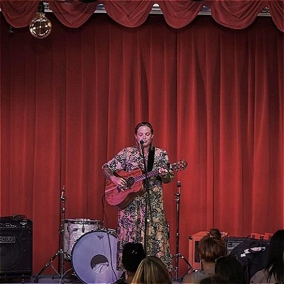 A performer in a patterned dress stands on stage in front of red curtains, playing a guitar