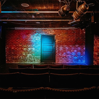 Image taken from the back of the venue. There are four rows of seats facing towards the performance space where the backdrop is a brick wall with a black door in the centre. Rainbow lighting illuminates the space.
