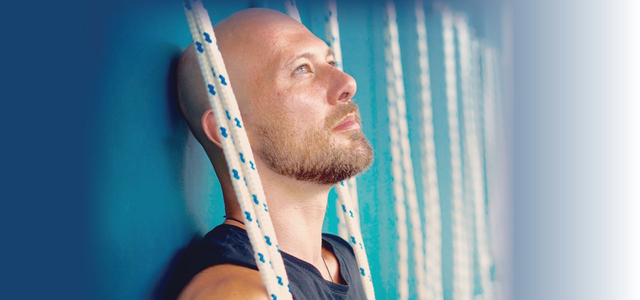 A person with tan skin and facial hair looking upwards while leaning on an ocean blue wall. White ropes hang over their shoulders.