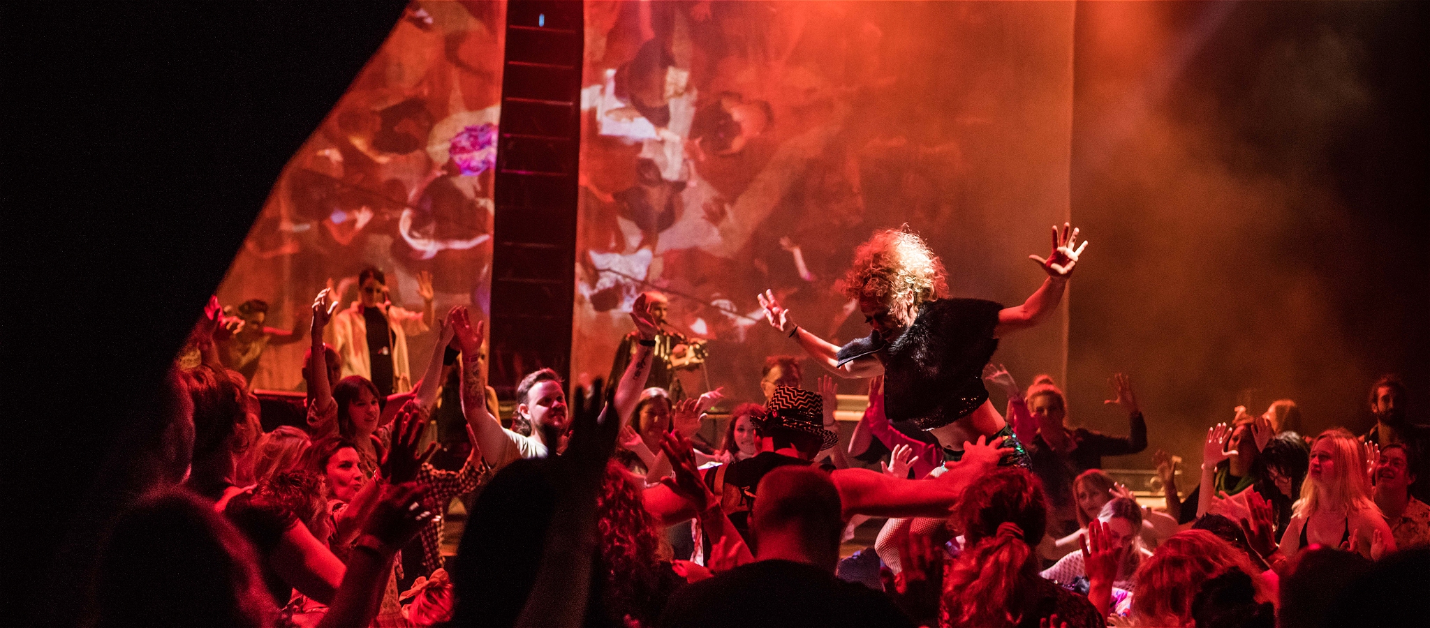A diva-like performer surrounded by a mass of humanity under red lighting