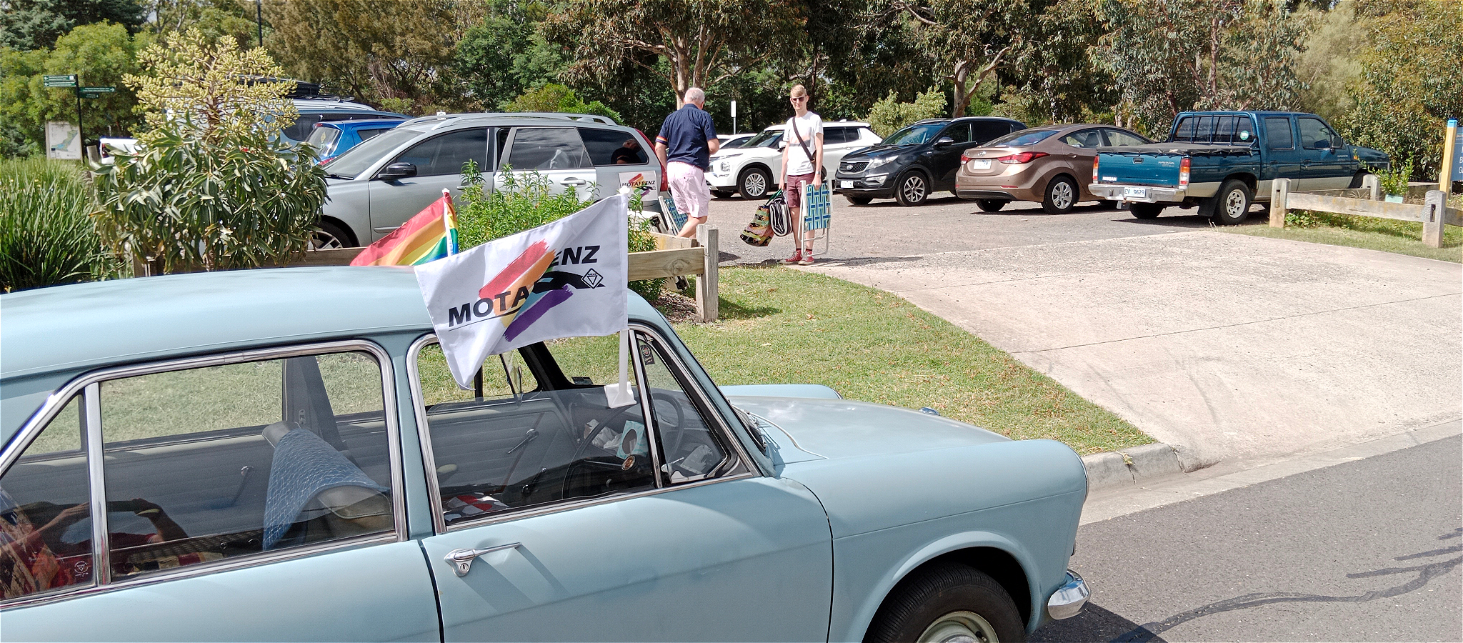A light blue vintage car with rainbow flags on the side, parked next to a car park.