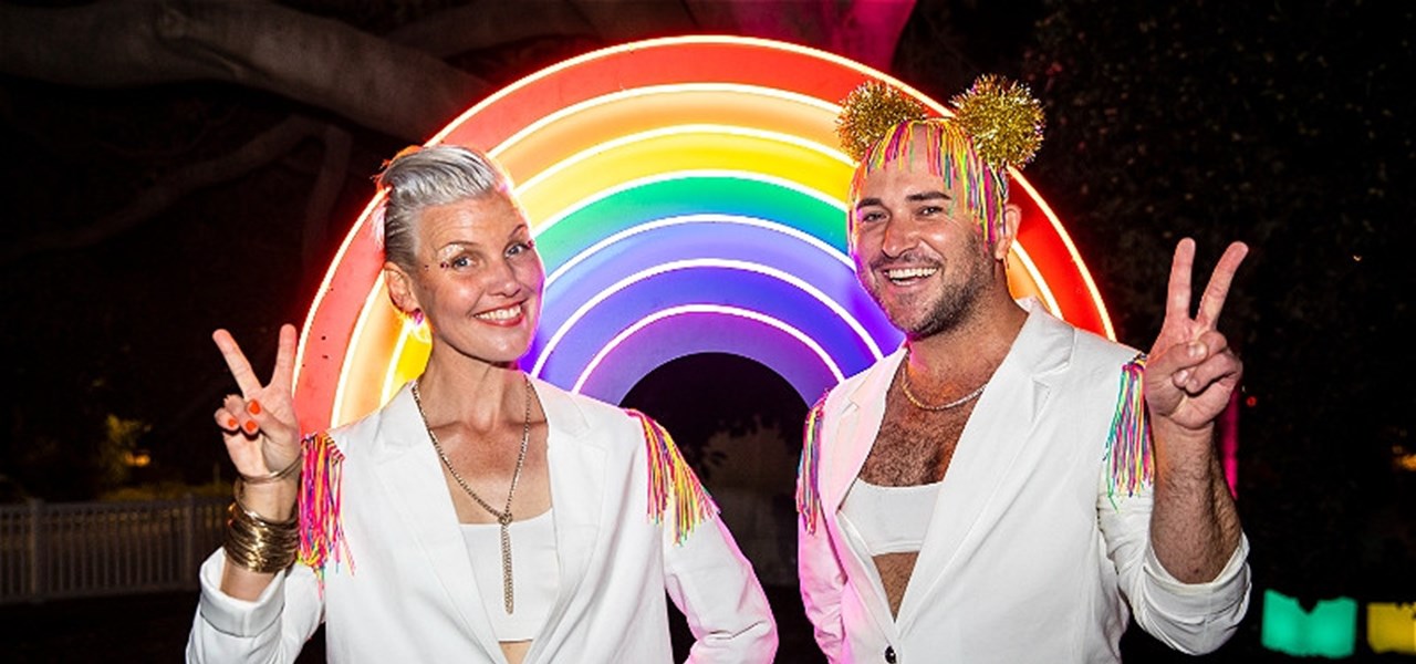 Bec Reid and Tristan Meecham dressed in matching white suits, looking happy in front of a large rainbow.
