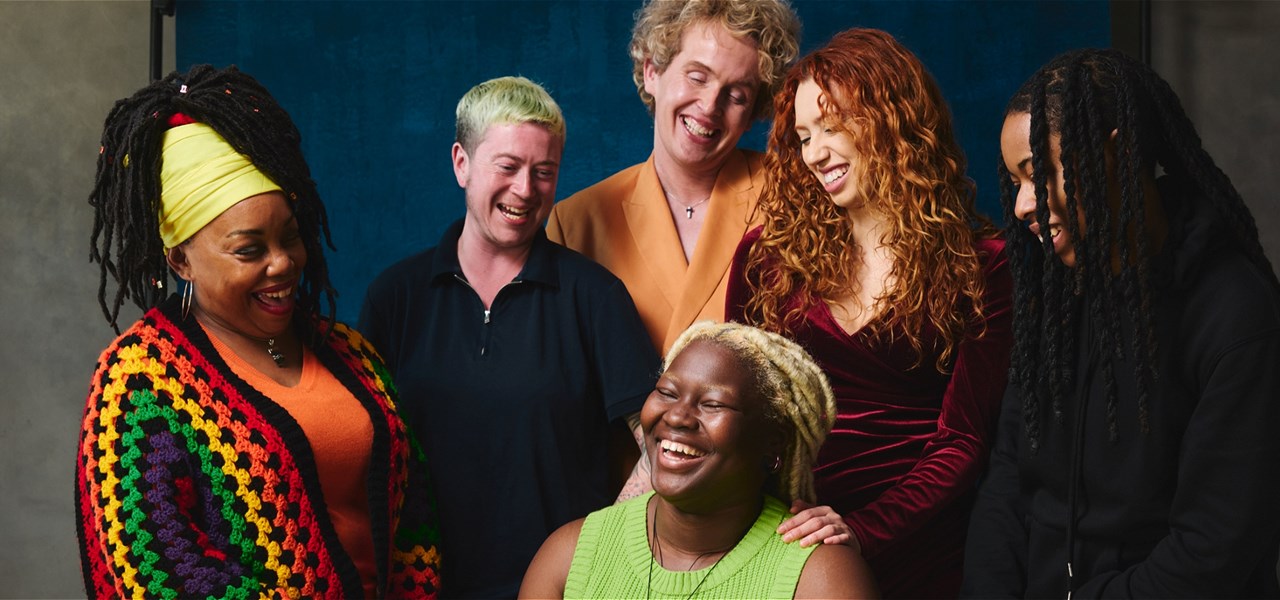 Portrait of six LGBTQIA queer people laughing against studio backdrop