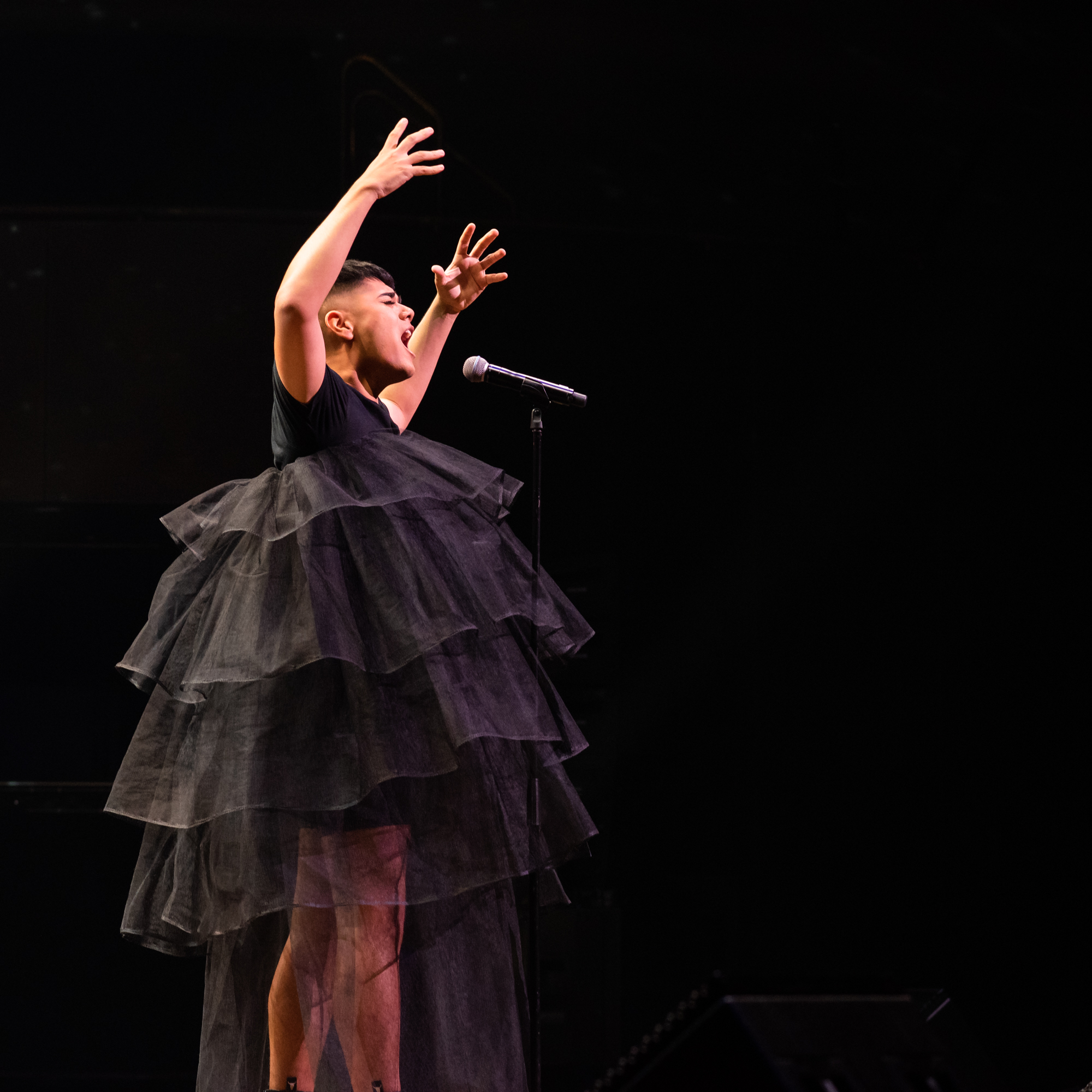 Person singing with arms outstretched upwards while singing and wearing a black tulle dress
