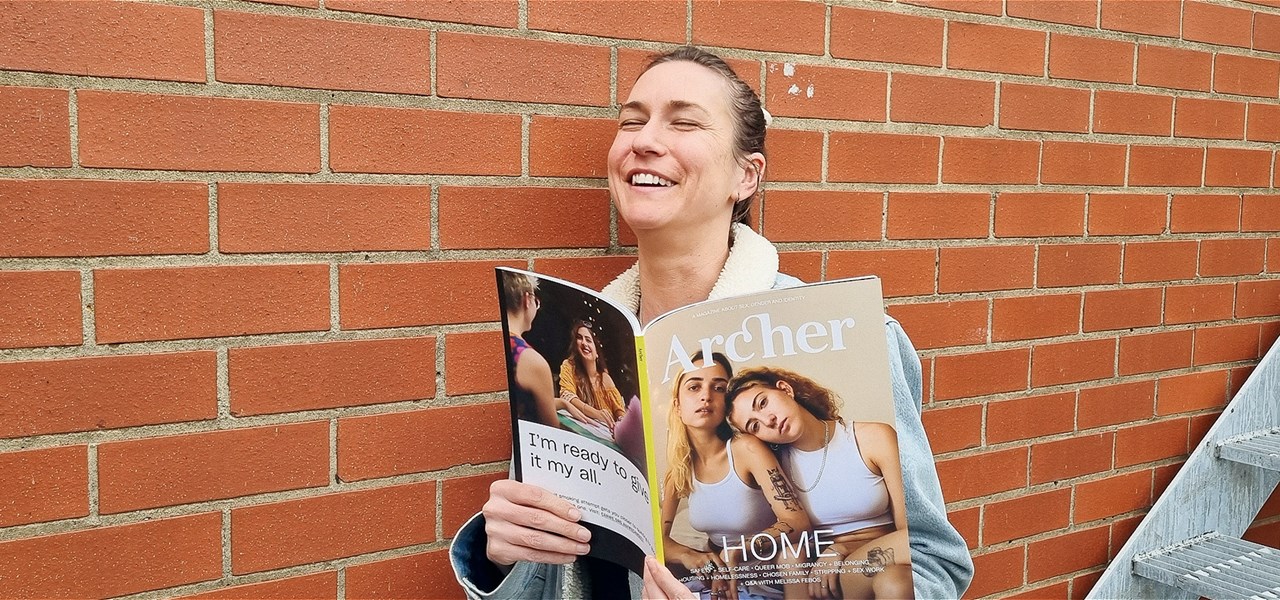 A happy looking person leafing through Archer Magazine, a plain brick wall behind them