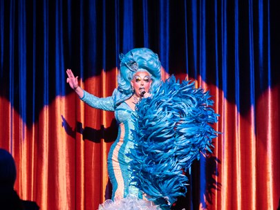 Abril La Trene on stage, in drag, dressed in a long blue dress with blue wig and large blue feather boa