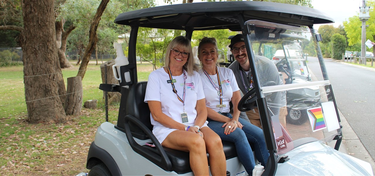 2 people wearing white t-shirts sit in a golf buggy and are smiling. Another person wearing a grey long-sleeved t-shirt is leaning into the golf buggy