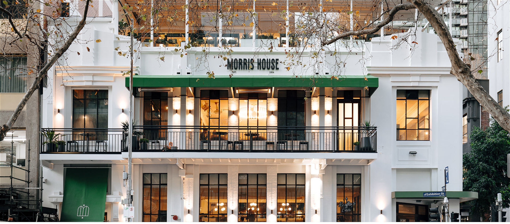 The white and green exterior facade of Morris House with warm light in the windows and framed by autumn trees on the street outside.