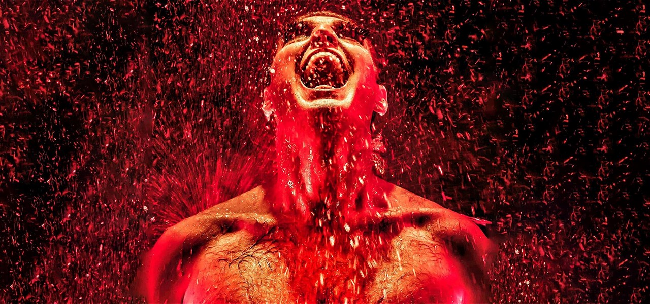 A bare-chested man with his mouth open, standing in the rain, and what appears to be fire and sparks pouring out of his mouth.
