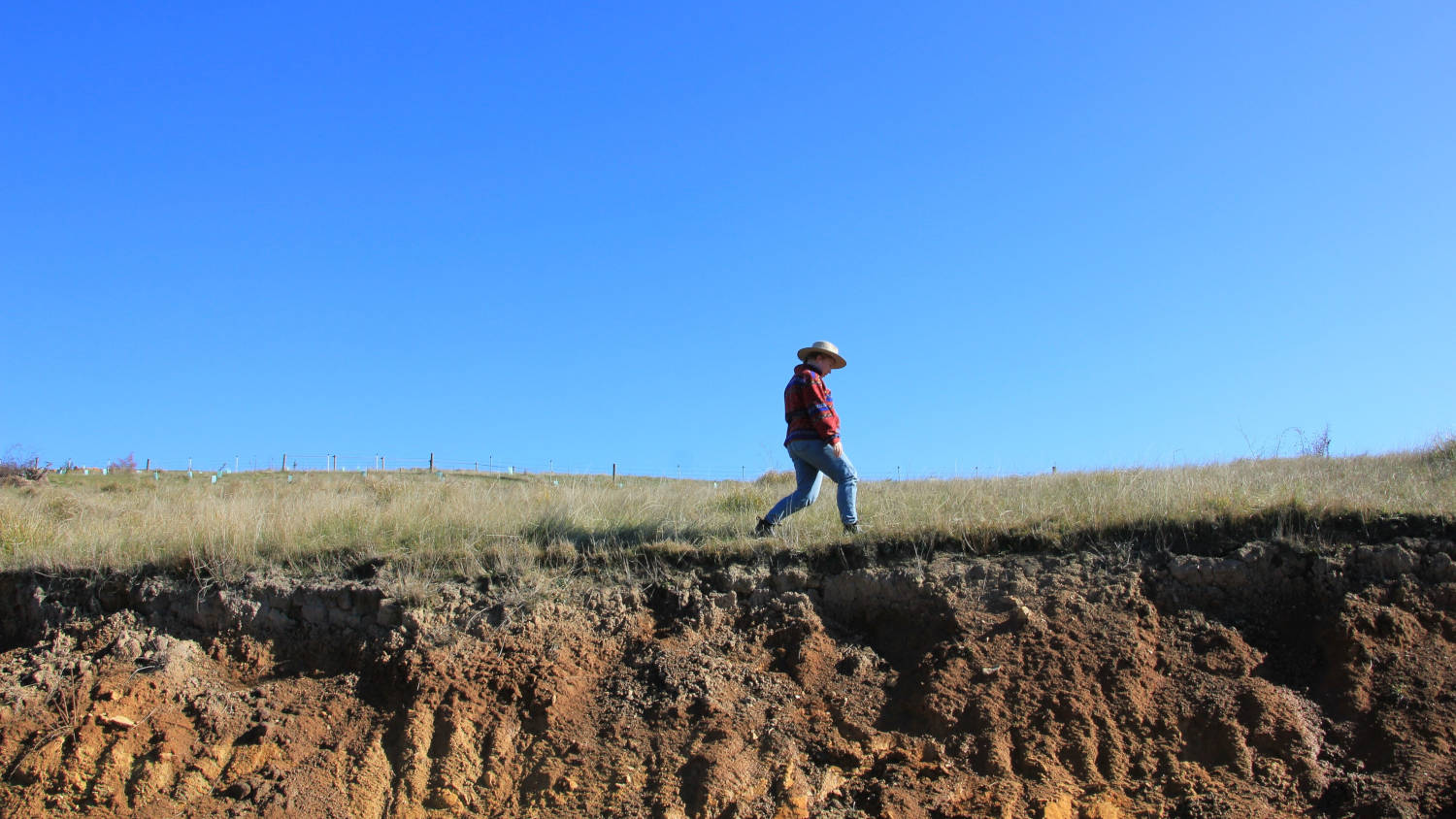 Lone figure of a person walking across a dry field, with eroded cliffs in the foreground and a bright blue sky