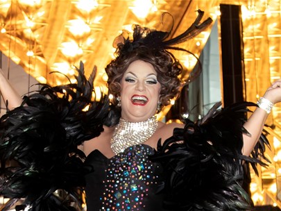 Dolly stands on the steps of a theatre. There are Lights and mirrors either side of her. She is wearing a black gown with sparkles, and smiling.