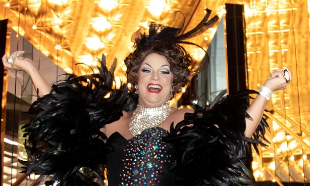 Dolly stands on the steps of a theatre. There are Lights and mirrors either side of her. She is wearing a black gown with sparkles, and smiling.