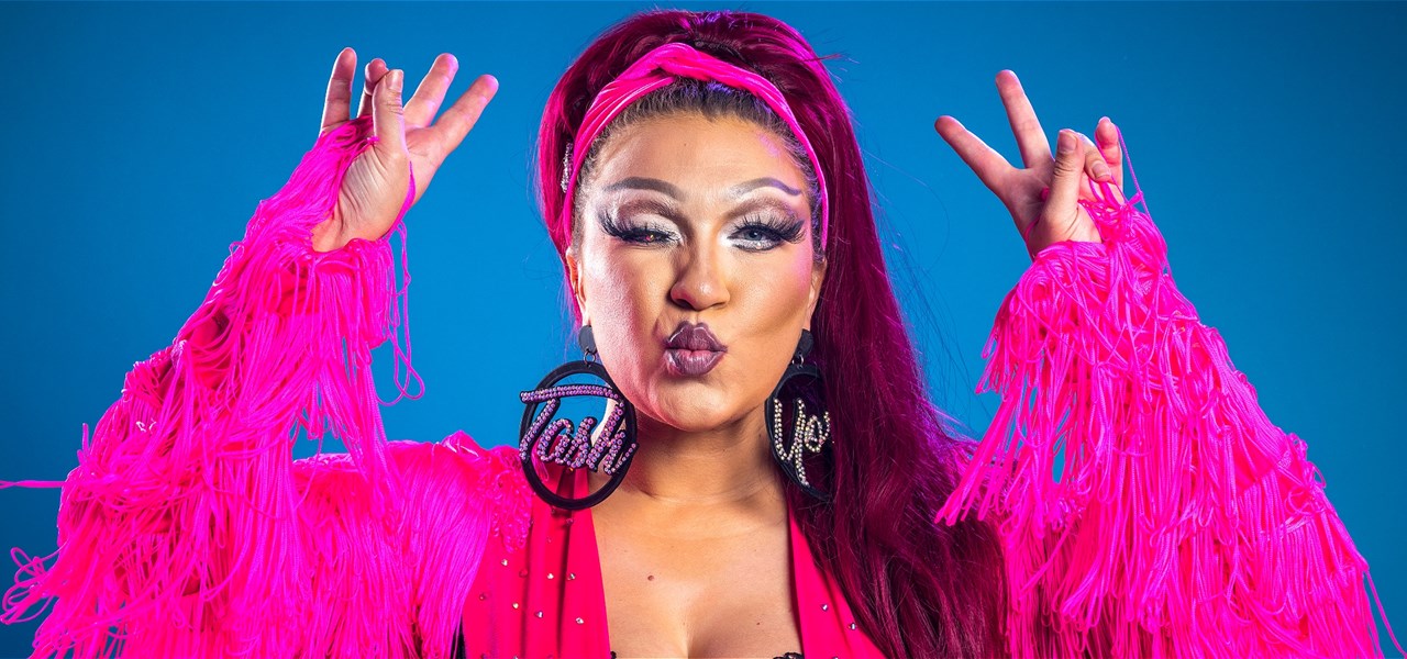 A woman wearing a hot pink tassled long sleeve shirt with a pink head band and oversized earrings. She is closing one eye and doing the peace fingers