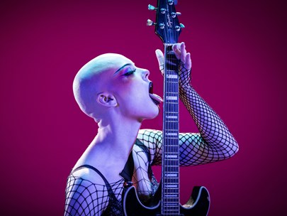 Person with a shaved head dressed stylishly in black lace licking the neck of an electric guitar
